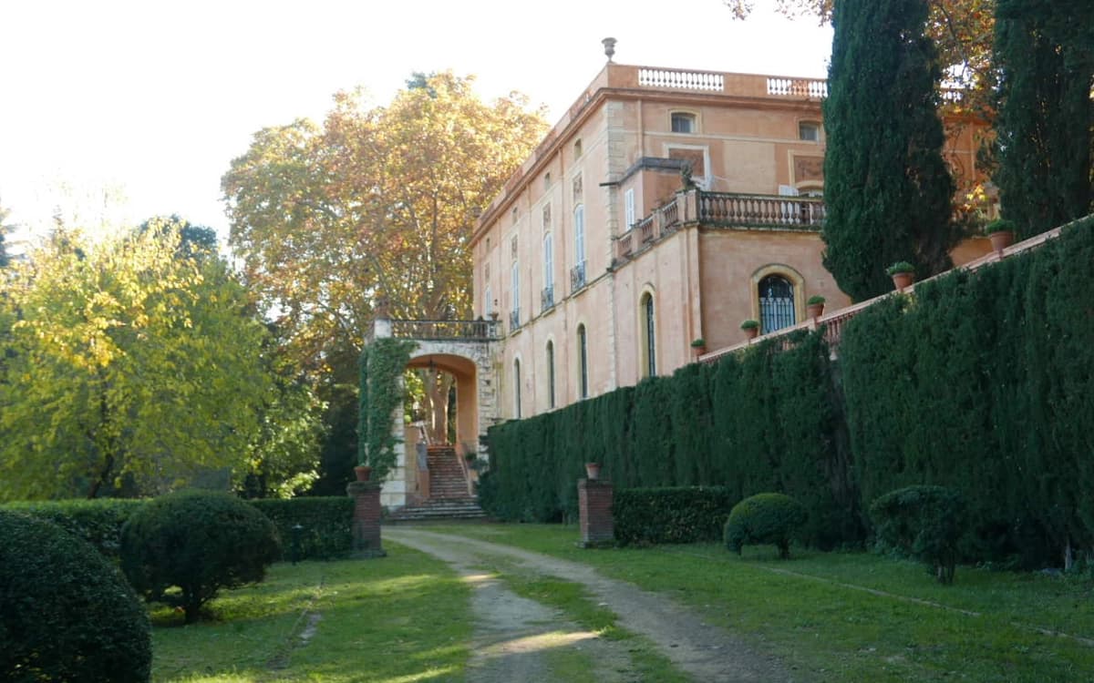 Historic palace garden in Barcelona's Horta-Guinardó area with tree-lined path, hedges, stone stairs, and warm exterior facade.