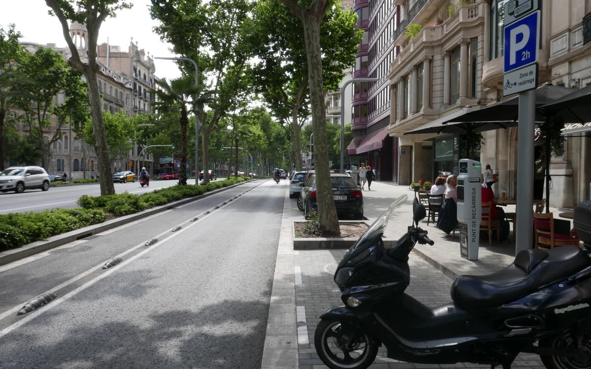 Passeig de Gràcia street in Barcelona with tree-lined boulevard, traffic lanes, sidewalk cafes, buildings, and parked scooter.
