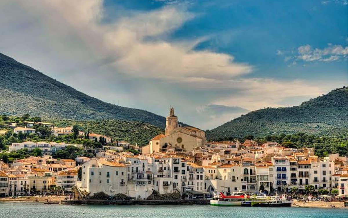 Whitewashed fishing village of Cadaqués in Girona with church tower, waterfront bay, boats, hills, and cloudy sky.