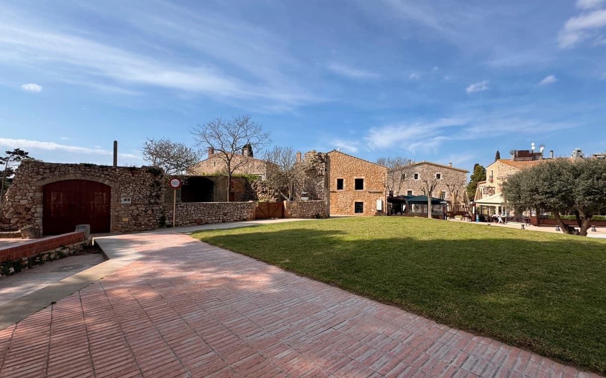 Stone houses around a village square in Pals, Girona, with lawn, brick walkway, trees, and blue sky.