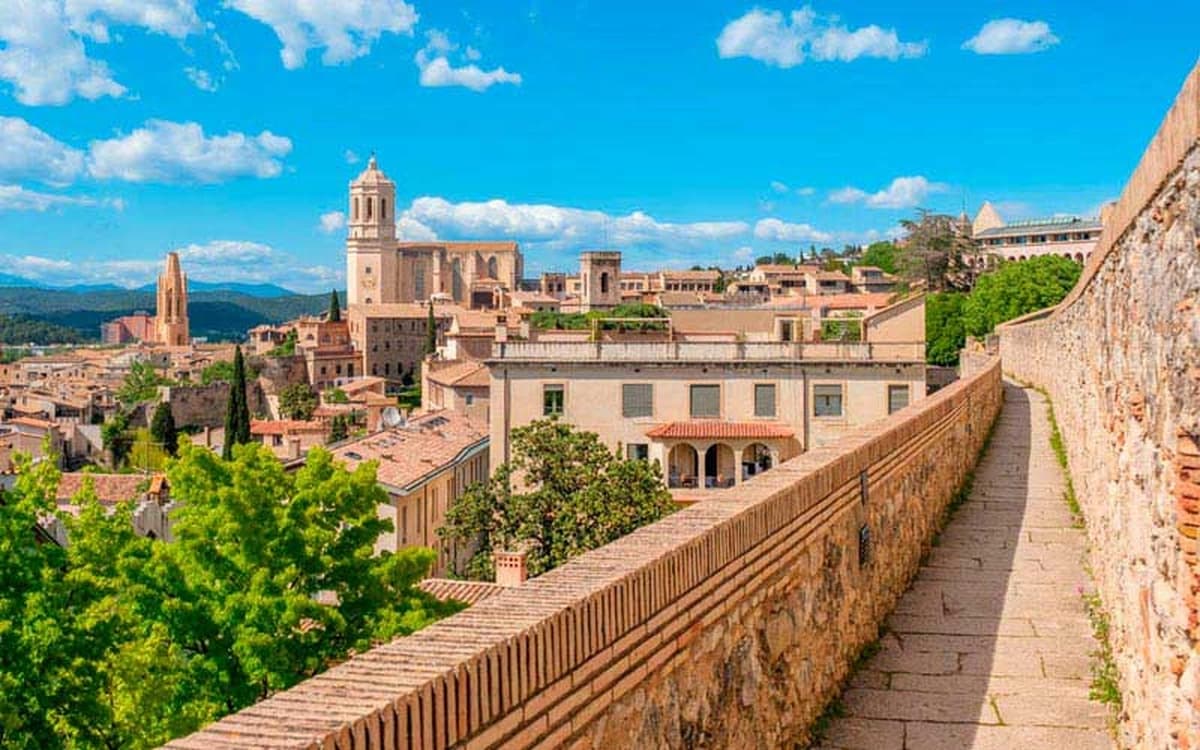 Girona old town skyline with medieval city walls, cathedral towers, stone walkway, rooftops, and green hills.