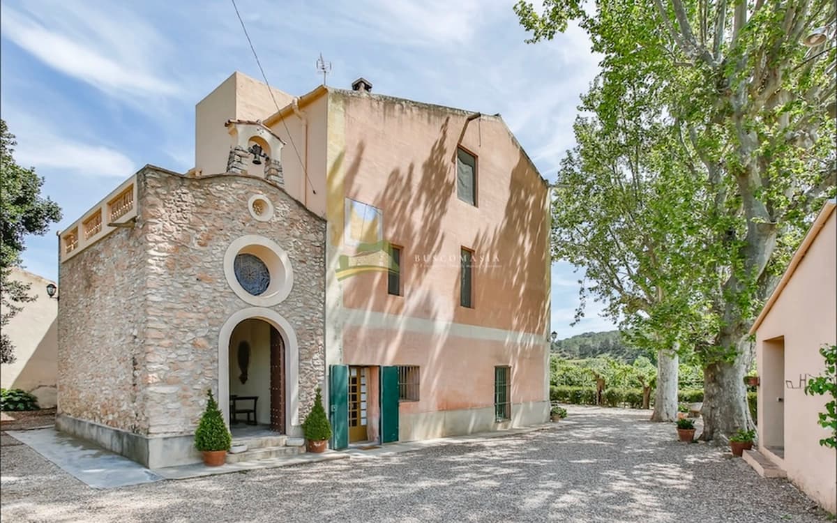 Cosido Farm chapel and masia courtyard in Tarragona with stone facade, pink farmhouse wall, gravel ground, trees, and open exterior space.