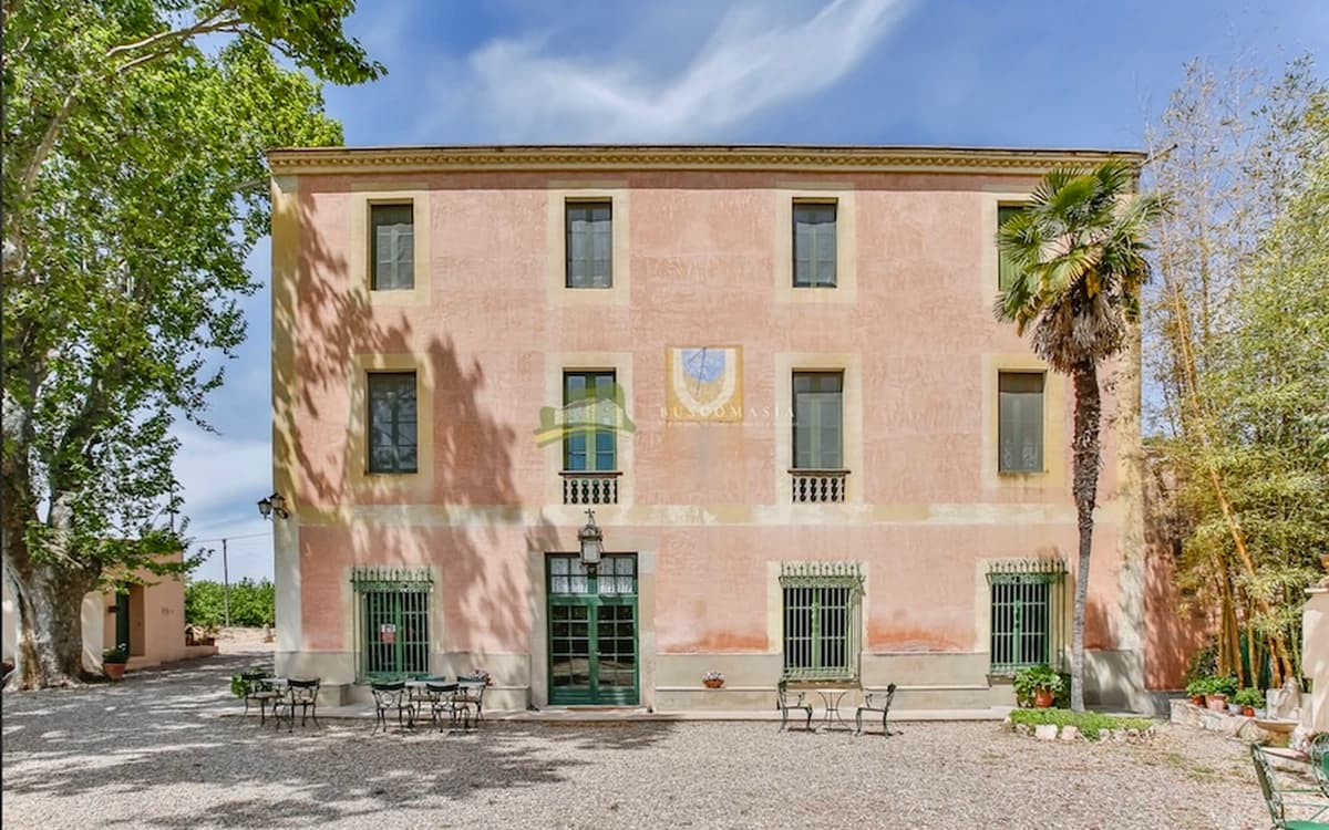 Cosido Farm masia facade in Tarragona with pink exterior walls, green doors, gravel courtyard, trees, and outdoor seating.