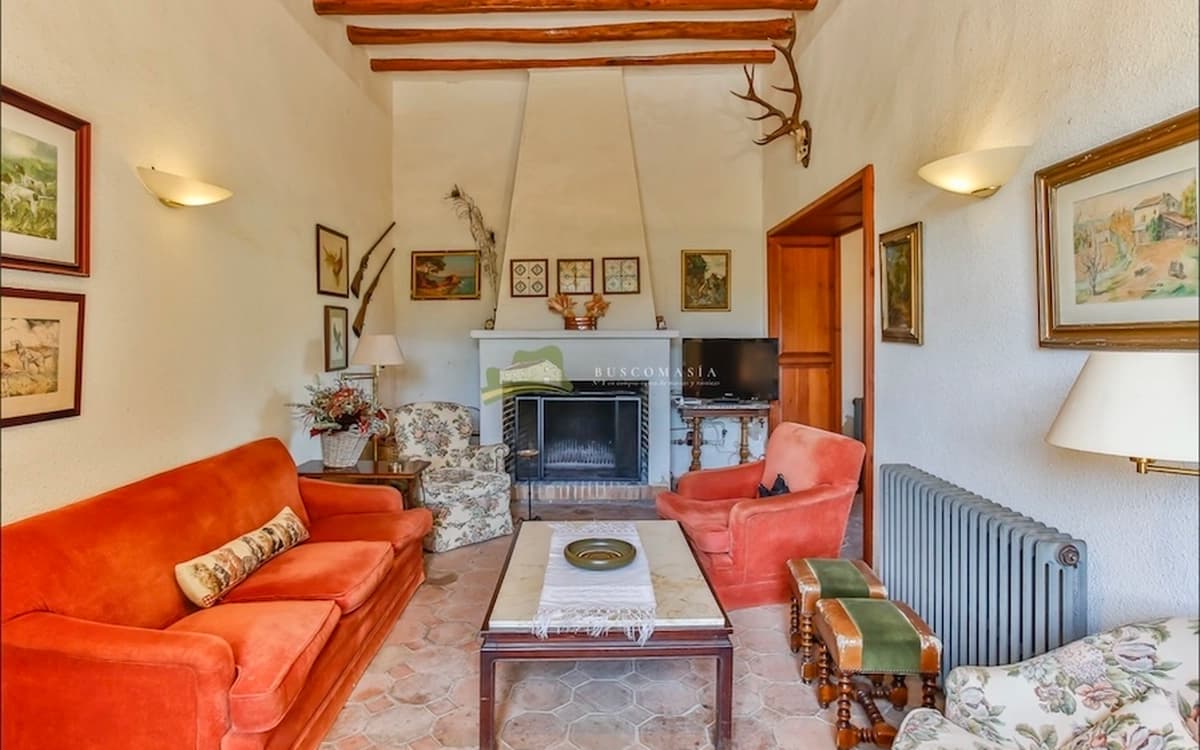 Rustic living room at Cosido Farm in Tarragona with fireplace, wooden beams, vintage seating, framed artwork, and warm interior texture.