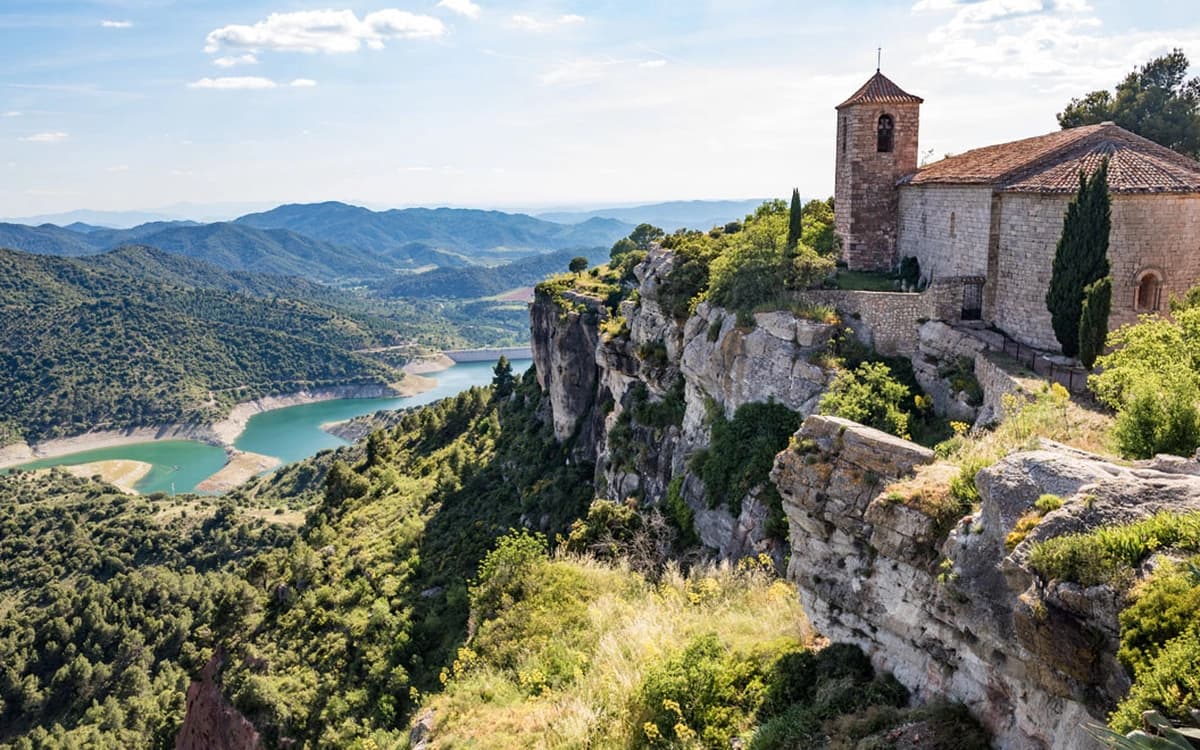 Cliffside church in Siurana near Tarragona overlooking a reservoir, forested valley, rocky cliffs, and mountain landscape.