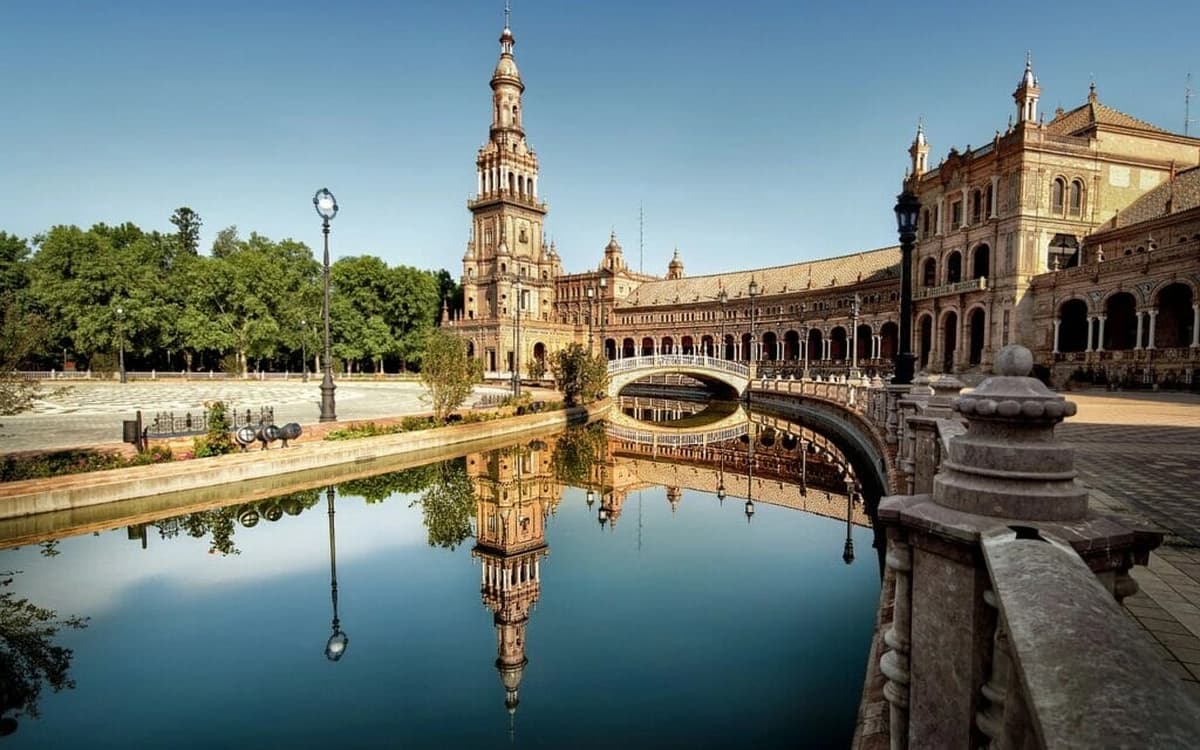 Plaza de España in Seville, Andalusia with historic palace architecture, canal bridge, water reflection, trees, and clear sky.