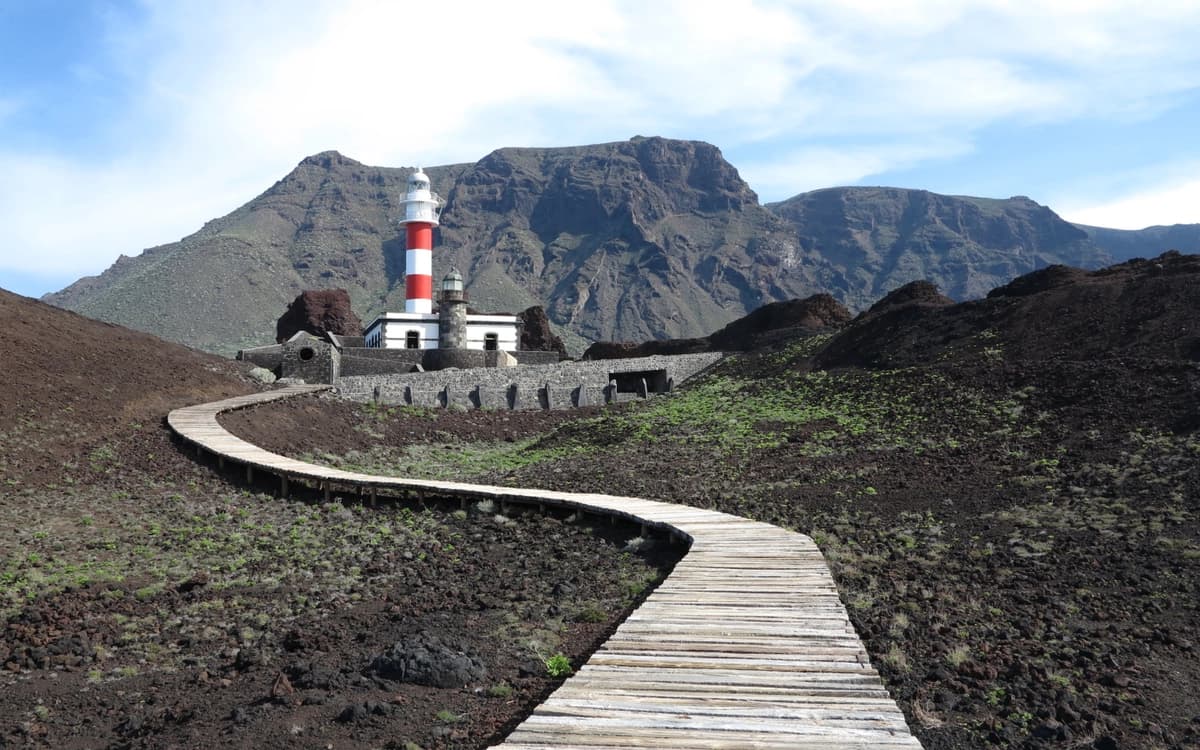Punta de Teno lighthouse in Tenerife with volcanic ground, wooden boardwalk, rugged mountains, and open sky.
