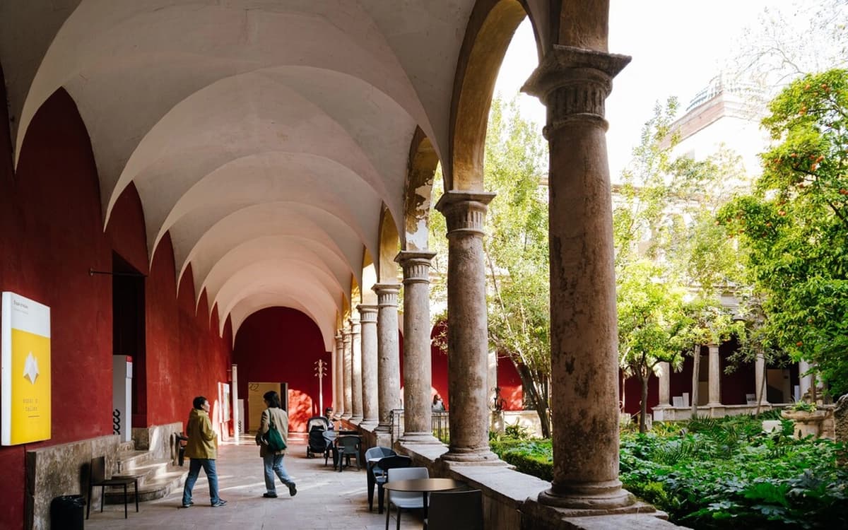 Historic cloister in Valencia's Barrio del Carmen with red walls, stone columns, arched walkway, garden courtyard, and visitors.