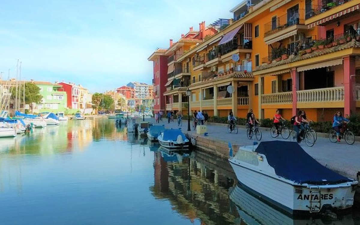 Port Saplaya marina canal near Valencia with colourful waterfront buildings, moored boats, promenade, cyclists, and calm water.