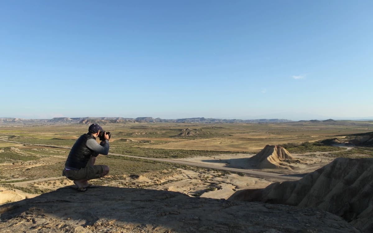 Location scout photographing the Bardenas Reales desert landscape with open plains, rock formations, roads, and clear sky.