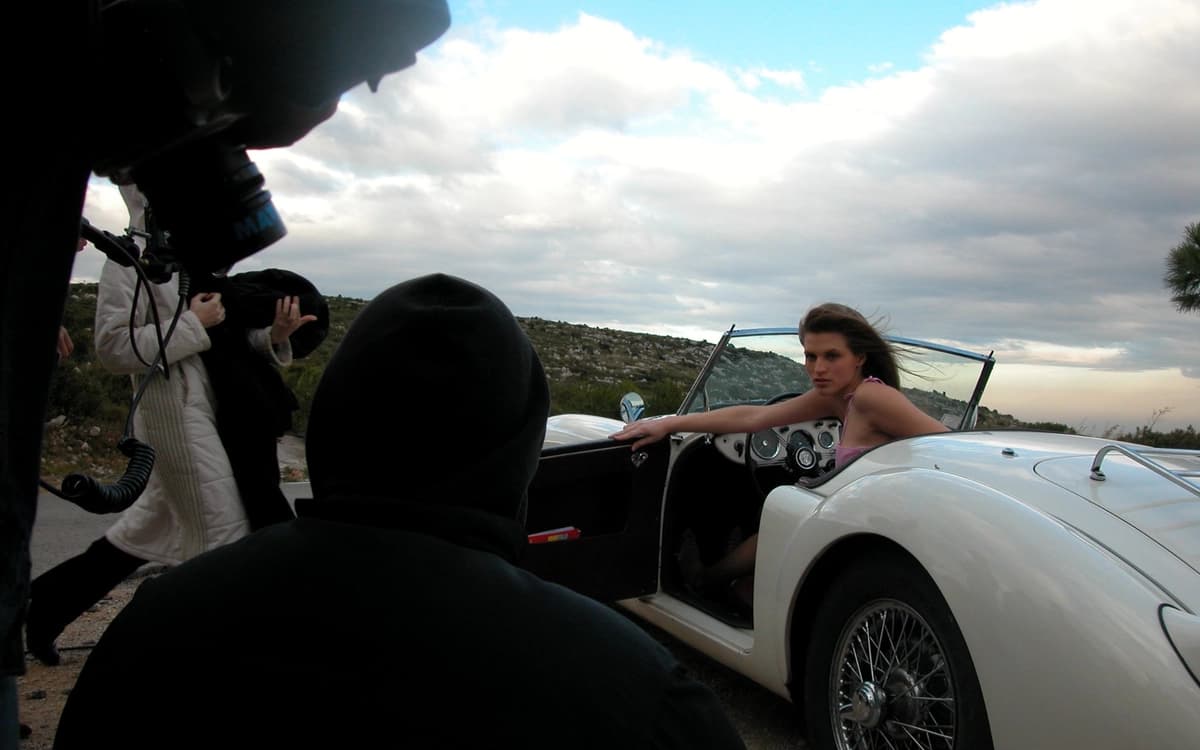 Crew filming a model in a white convertible on a roadside location with camera team, coastal hills, and cloudy sky.