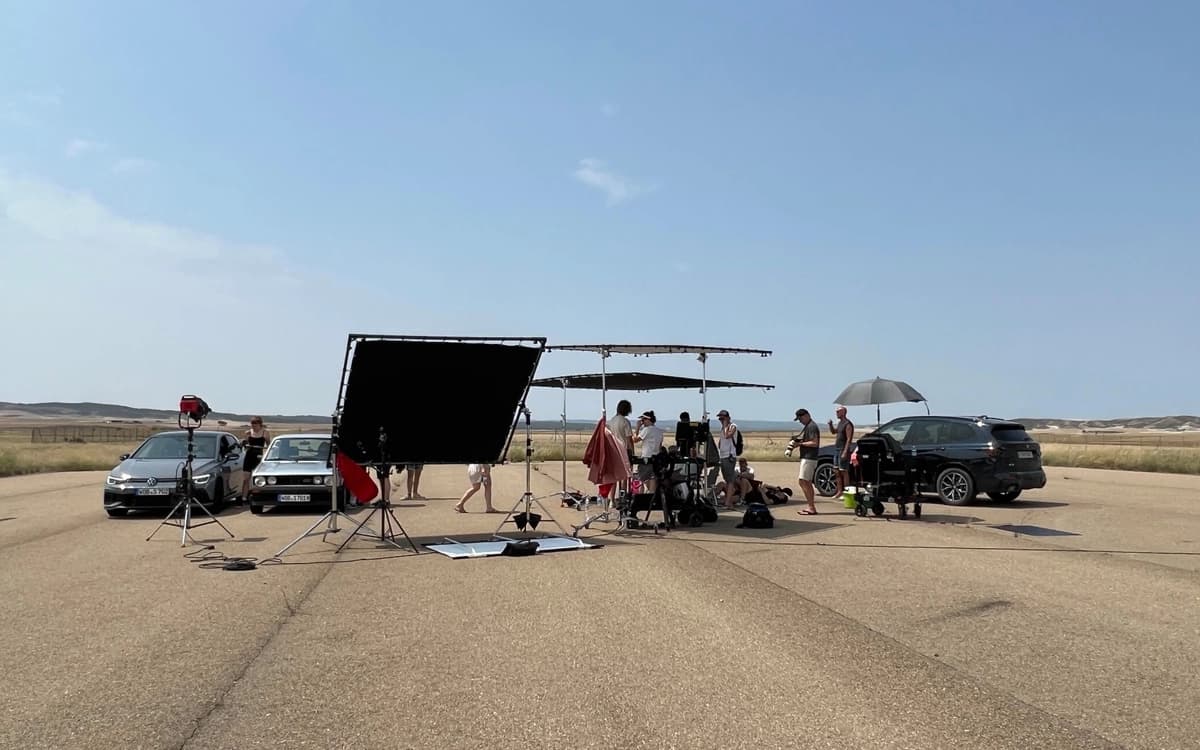 Production crew and vehicles set up on a wide desert highway with lighting frames, shade, camera equipment, and open terrain.