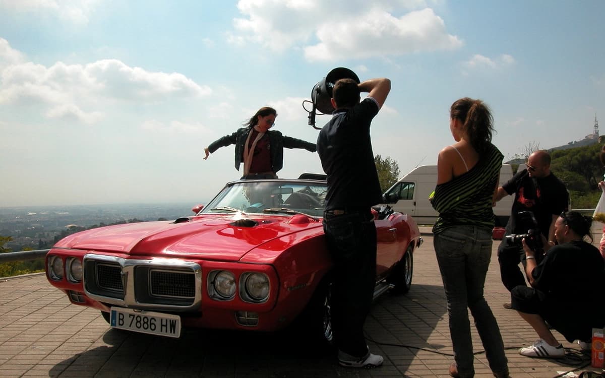 Production crew filming talent in a red classic car on a roadside location with camera, lighting, and support team.