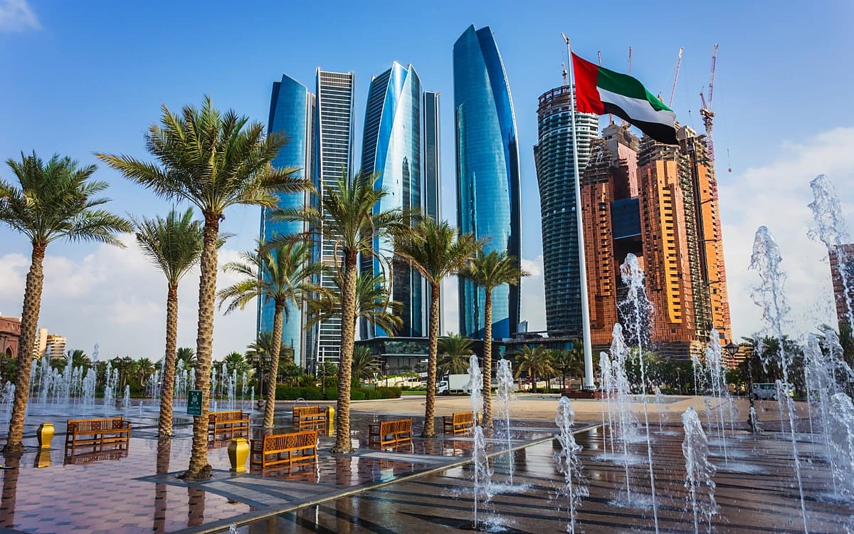 Abu Dhabi skyline with modern towers, palm trees, fountains, UAE flag, and city waterfront setting.