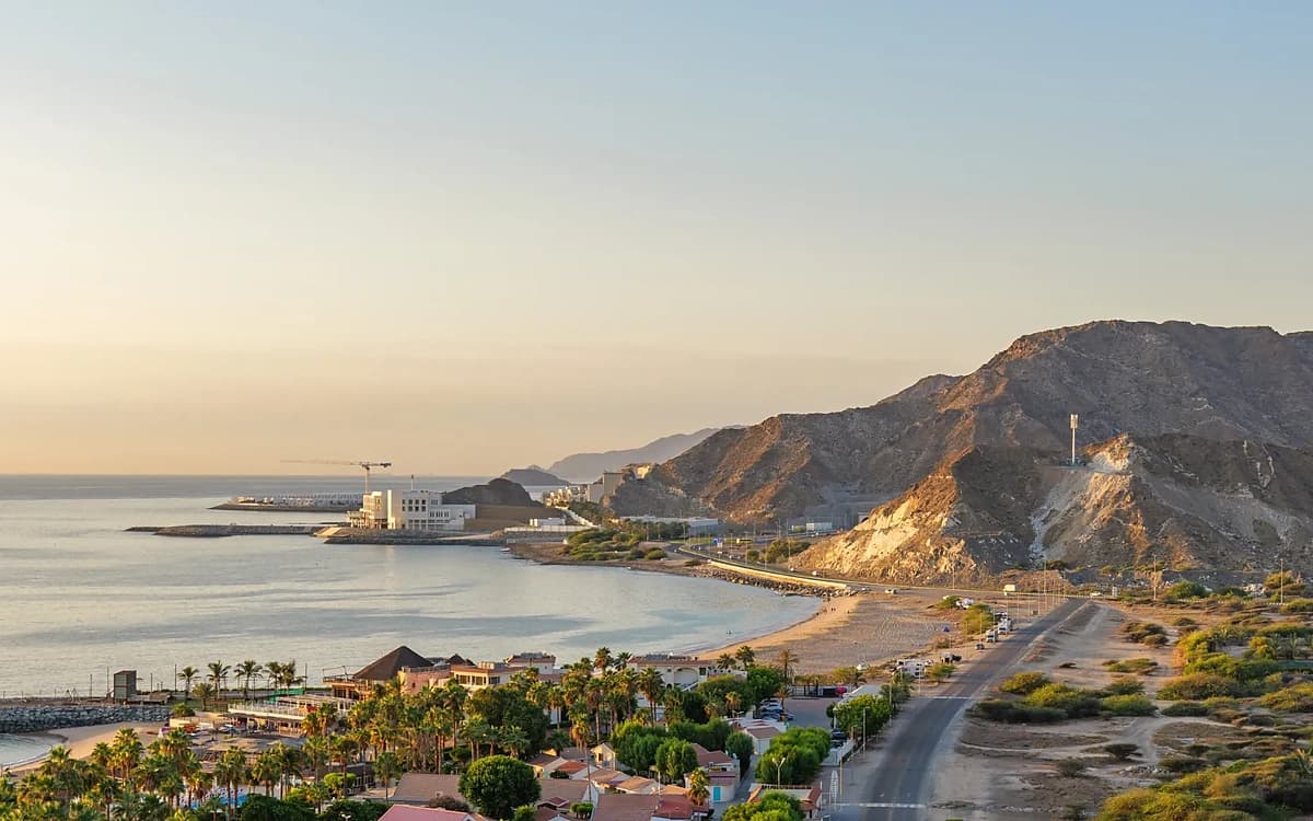 Fujairah coastline with beach, mountain road, coastal buildings, and Gulf of Oman water at golden hour.