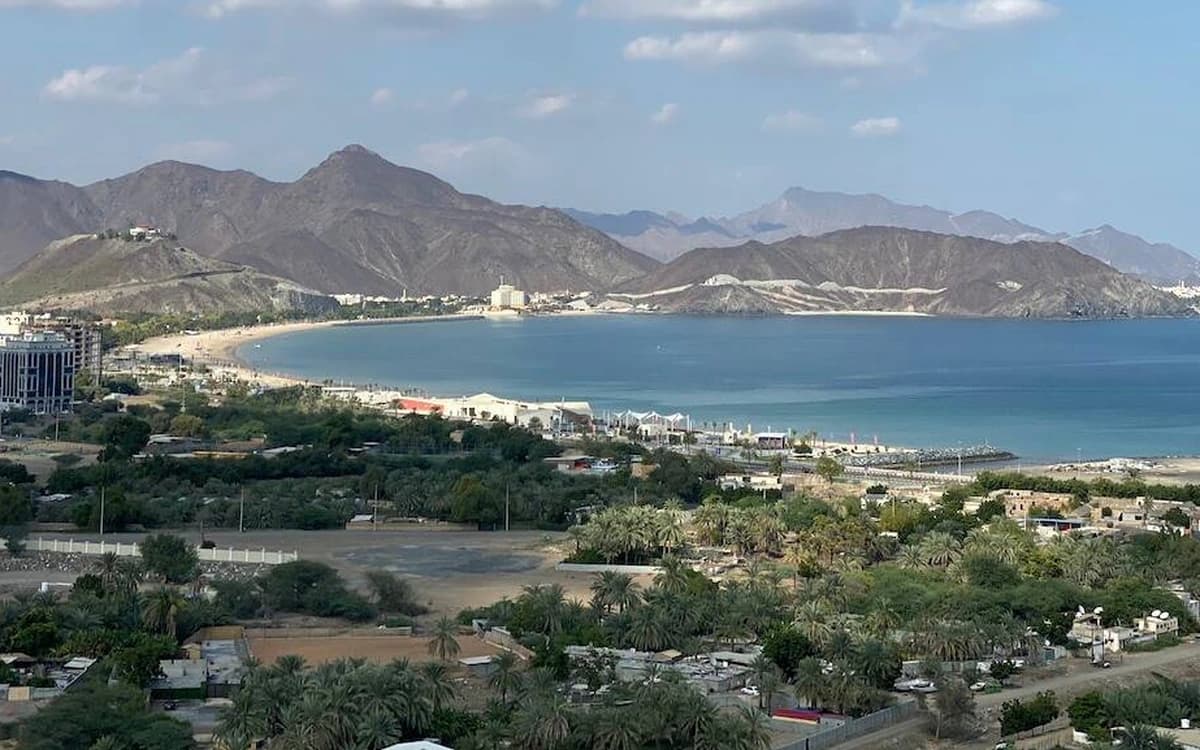 Mountain coast in the UAE with blue Gulf water, coastal town, palm trees, and Hajar mountain backdrop.