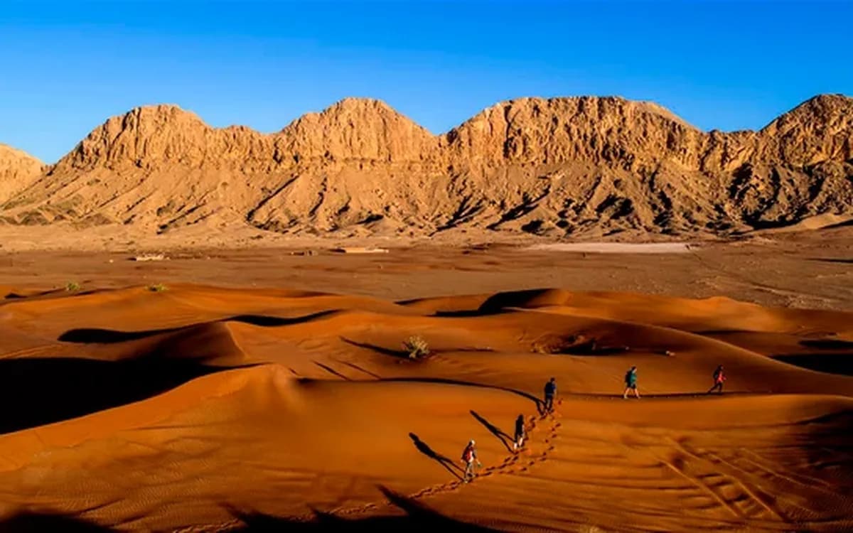 Desert landscape in Sharjah with red dunes, rocky Hajar mountain backdrop, and people crossing open sand terrain.