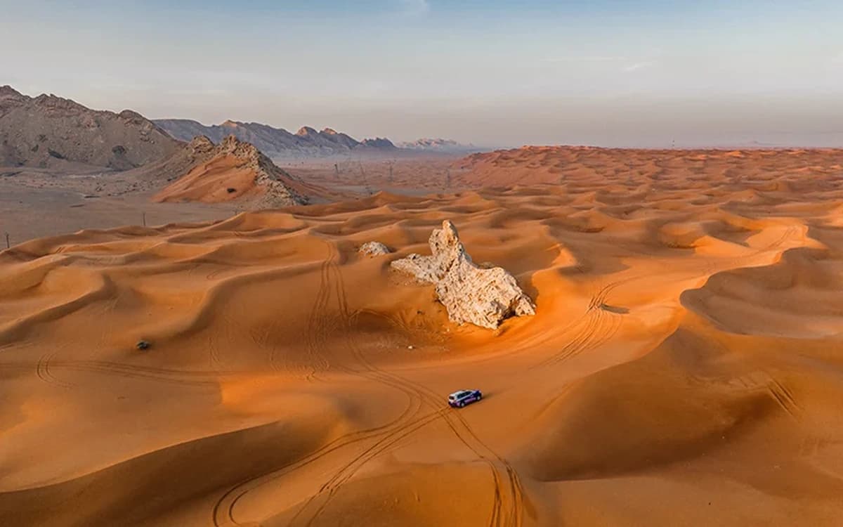 Desert dunes and pale rock formations in Sharjah with mountain backdrop, vehicle tracks, and open archaeological desert terrain.