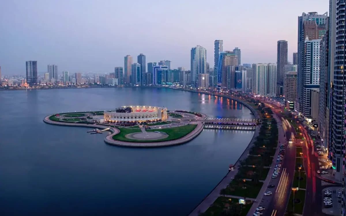 Sharjah lagoon waterfront with city skyline, curved coastal road, island landmark, and evening light.