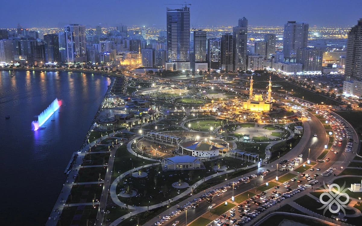 Sharjah waterfront skyline at night with lagoon, city towers, illuminated mosque, roads, and public parkland.
