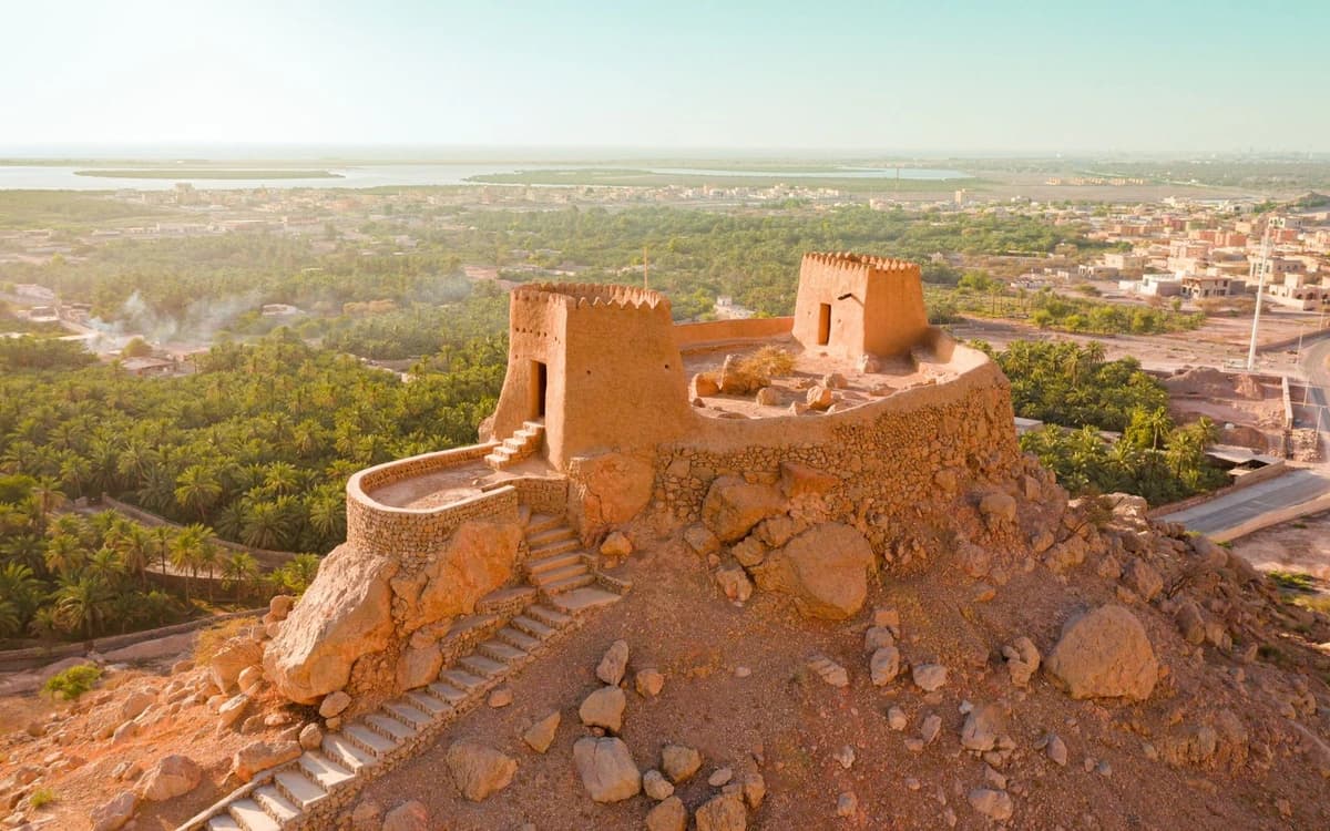 Hilltop fort structure in Ras Al Khaimah with stone walls, palm oasis, coastal water, and surrounding town.