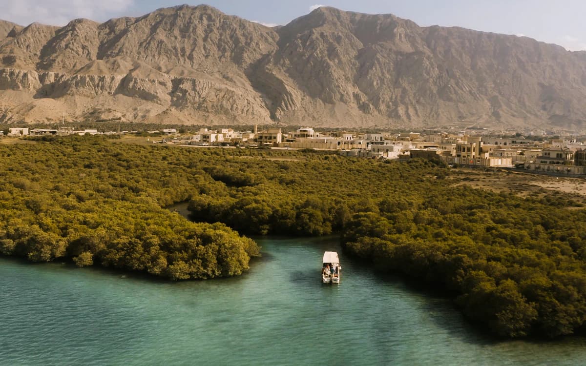 Ras Al Khaimah mangrove-lined water channel with small boat, turquoise water, coastal town, and rocky mountains behind.