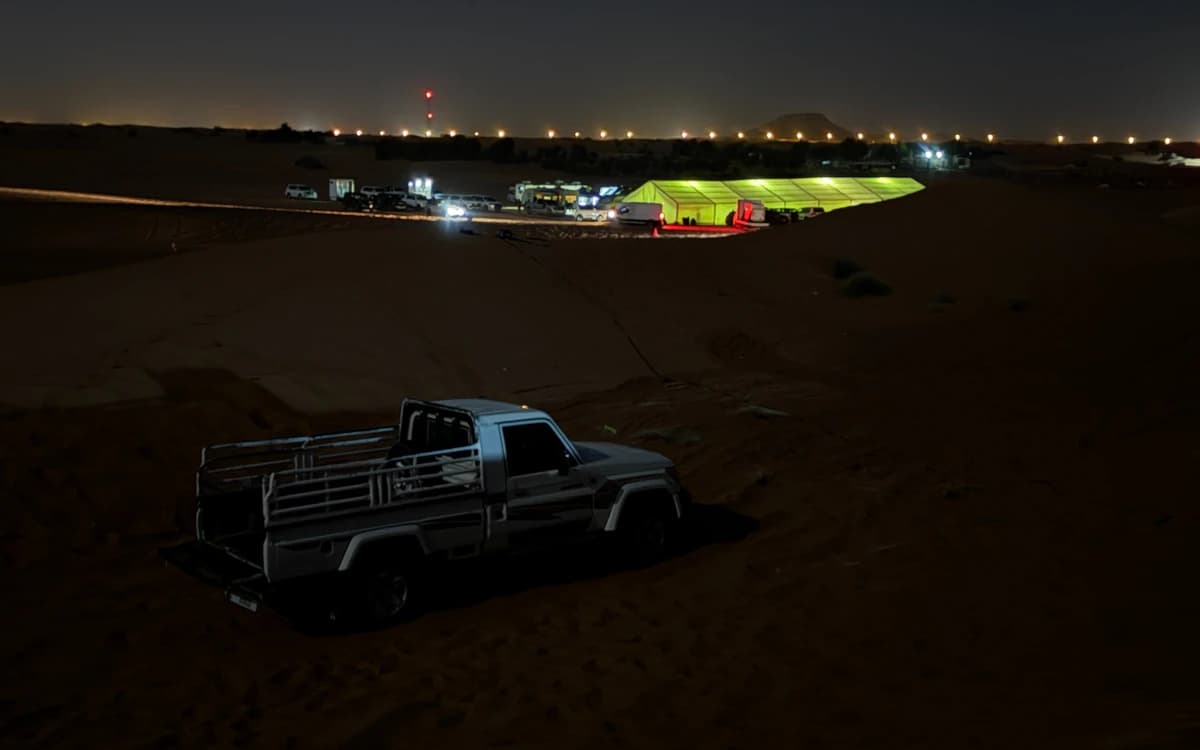 Night desert production logistics setup in the UAE with vehicles, lit unit tent, crew base area, cables, and sand dunes.