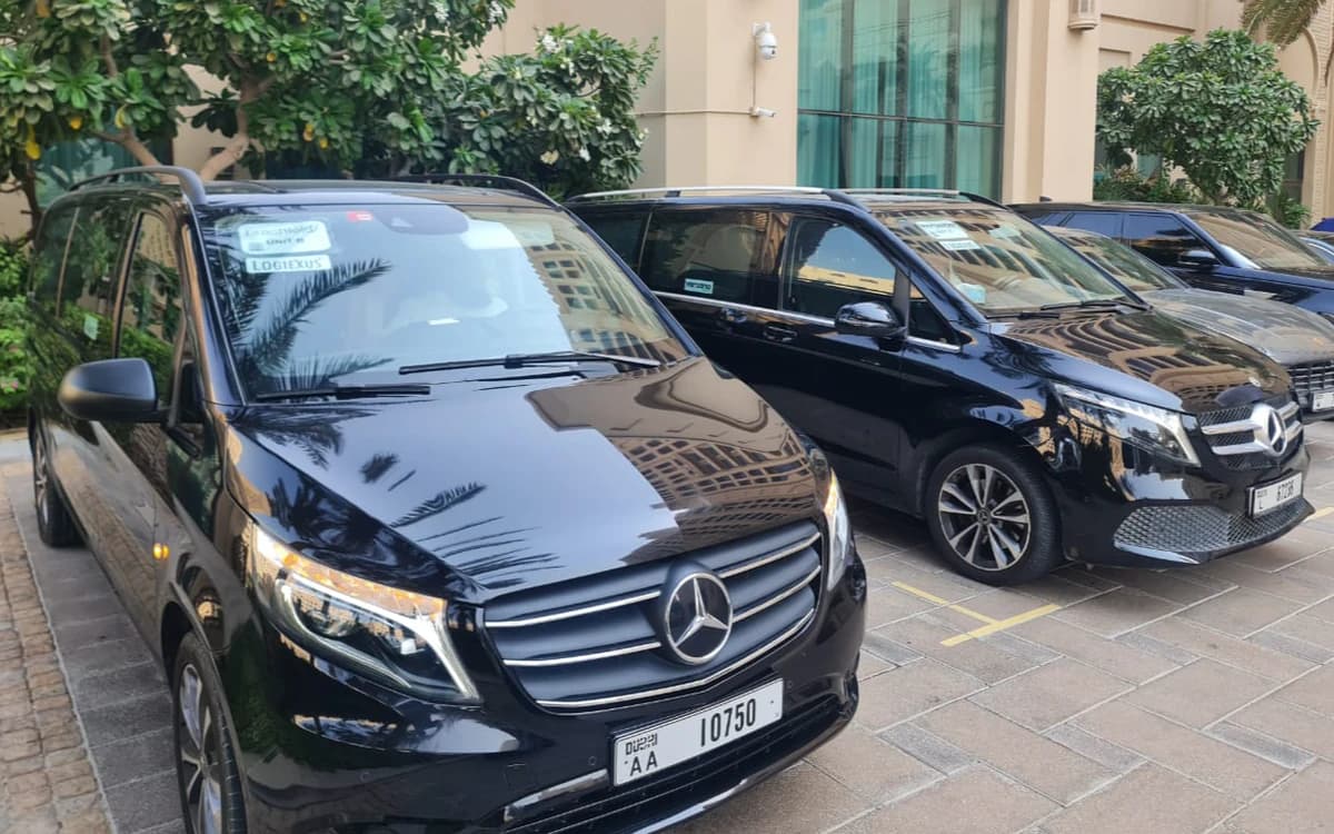 Production transport vehicles in the UAE parked beside a building with greenery, hotel-style exterior, and logistics stickers.